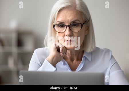 Serious older age woman in glasses office worker ceo focused at work on computer screen solve business problem. Busy aged female lawyer think on contr Stock Photo