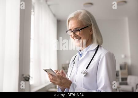 Smiling older woman doctor using mobile phone at work typing referral at digital app writing message to patient check record at medical database. Happ Stock Photo
