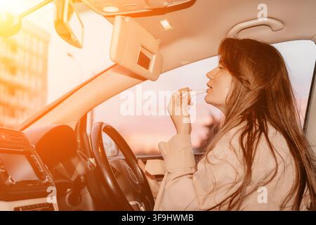 Woman lipstick car applying makeup in driver seat using sun visor mirror during a beautiful golden sunset flare Stock Photo