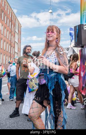 COPENHAGEN, DENMARK - AUGUST 15, 2026: Portrait of a young female participant in a creative festival costume during the annual Copenhagen Pride parade in Copenhagen, Denmark. The woman is styled with pink rimless sunglasses, a metallic sequin vest, a holographic fanny pack, and rainbow-colored fishnet tights. She holds a smartphone and a beverage can while standing in the street next to a parade sound truck. In the background, traditional brick apartment buildings and other spectators fill the festive scene celebrating LGBTQ rights, diversity, and equality. Editorial Only. Credit: Stig Alenäs Stock Photo