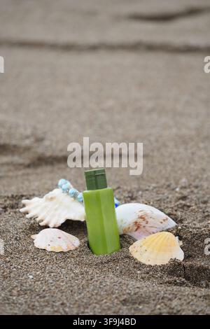 a small green plastic bottle in the sand, surrounded by several white and yellow seashell on a beach Stock Photo