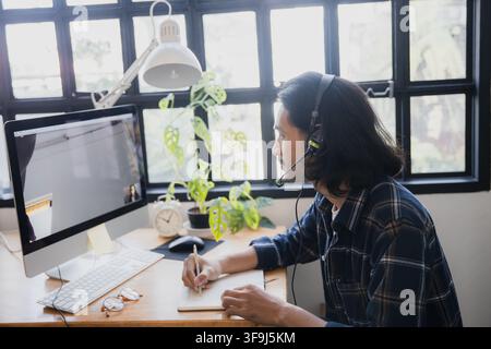 Young man wearing headset taking notes during online meeting at home office desk Stock Photo