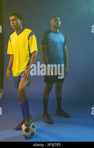 Two male athletes posing on blue seamless backdrop, wearing soccer jerseys and balancing ball Stock Photo