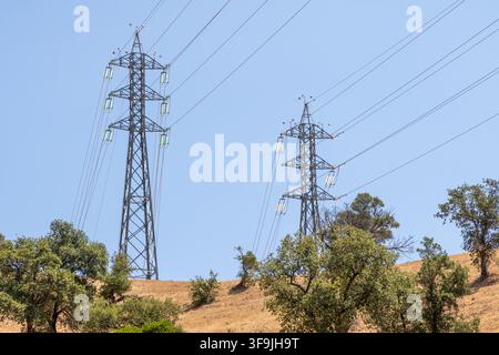 Two large metal electricity pylons stand tall against a clear blue sky, with power lines stretching between them over a sun-baked landscape Stock Photo
