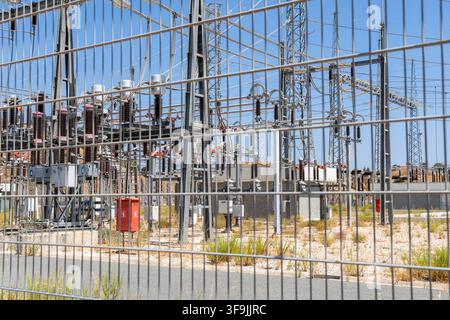 A view through a wire fence of a power substation featuring transformers, insulators, and transmission towers under a clear blue sky Stock Photo