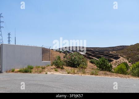 A large-scale solar panel array stretches across a parched, rolling landscape, with power lines visible in the distance and a concrete wall in the for Stock Photo
