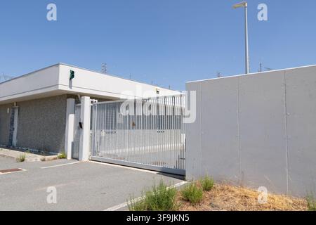 A modern industrial facility entrance featuring a tall metal security gate and a large concrete barrier under a clear blue sky Stock Photo