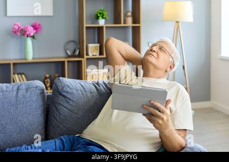 Elderly man with tablet on couch at home, stretching neck and relaxing indoors Stock Photo
