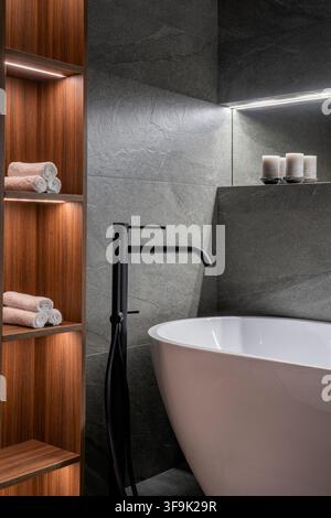 Vertical shot of freestanding bathtub with black faucet and wood shelving niche, luxury modern bathroom interior with folded towels Stock Photo