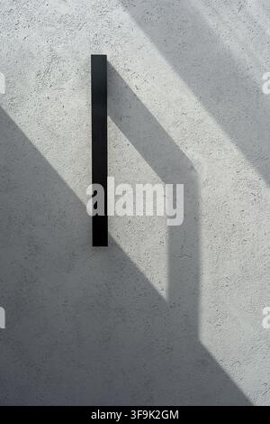 Minimalist black wall lamp mounted on a textured white plaster wall, creating dramatic geometric shadows and contemporary architectural lighting Stock Photo