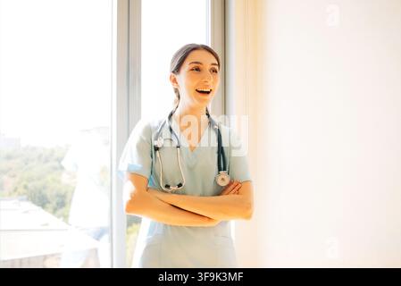 Female healthcare worker in light blue scrubs stands with arms crossed near a window, smiling in a bright and modern medical office setting Stock Photo