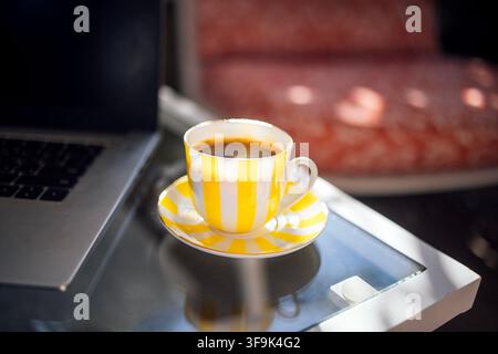 Yellow striped cup filled with coffee sits on a saucer beside a laptop on a glass table, with a softly blurred background of a comfortable seating are Stock Photo