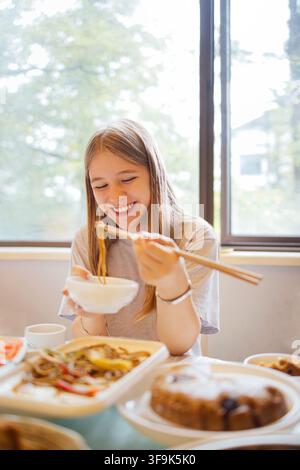 Young woman with long hair enjoys eating noodles with chopsticks, seated at a table filled with Asian cuisine, illuminated by natural light from a win Stock Photo
