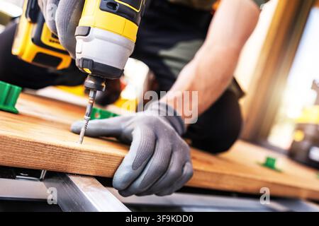 A person is focused on drilling a wooden board in a workshop. They wear gloves and use a cordless drill. Tools and materials are visible around them a Stock Photo