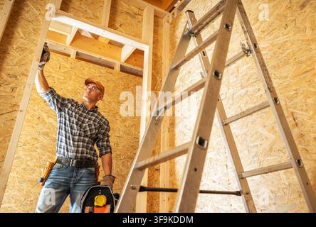 A man is working inside a building that is being renovated. He is using a measuring tool to check a wall. A ladder is nearby as he focuses on his task Stock Photo