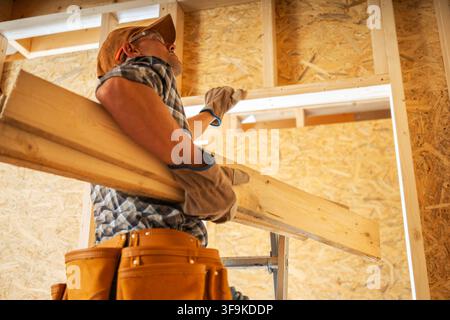 A worker is carrying a piece of wood inside a house that is being renovated. The scene shows the worker focused on the task. The walls are made of ori Stock Photo