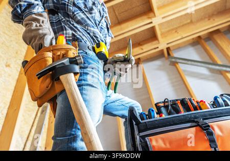 A construction worker stands in a workshop, wearing gloves and jeans. He holds a tool while a tool belt hangs at his waist. A tool bag is open nearby, Stock Photo