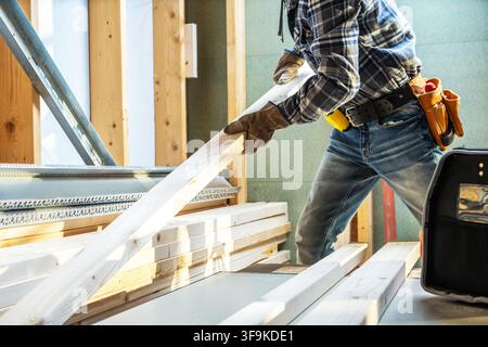 A person is carrying wooden boards in a construction site. The site has exposed wooden structures and tools are present. The worker wears gloves and a Stock Photo