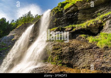 French Alps. Water flows down from high rocks in a forest during daytime. Sunlight shines on the waterfall and the surrounding greenery. The sound of Stock Photo