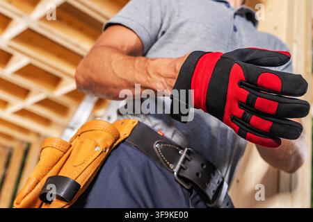 A person is getting ready to put on gloves while standing in a building site. The worker has a tool belt around the waist. The construction area shows Stock Photo