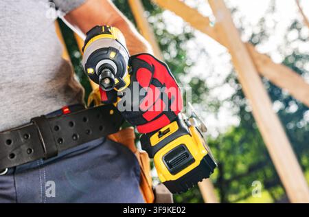 A worker stands at a construction site holding a power tool with both hands. The background shows wooden beams and greenery. The sun shines on the sce Stock Photo