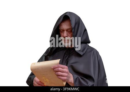 An elderly monk in a black hooded robe looks down while reading a parchment sheet isolated on a white background. Stock Photo