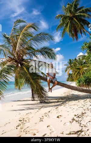 A man sits on a leaning palm tree along a tropical beach in Maldives Stock Photo