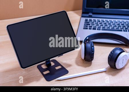 A tablet is on a stand next to a laptop and a pair of headphones. The tablet is black and the laptop is silver Stock Photo