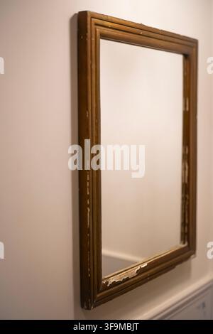 Rectangular vintage mirror with a distressed wooden frame hanging on a white interior wall, reflecting bright light in a room. Stock Photo