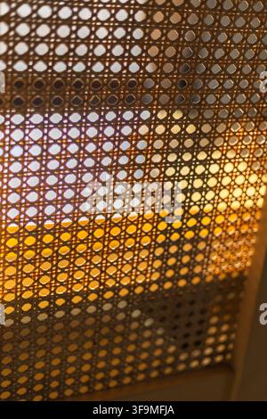 Detailed close up of natural woven rattan cane webbing texture backlit by warm sunlight on a wooden chair backrest in an interior setting. Stock Photo