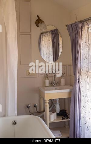 Bathroom interior featuring a ceramic washbasin vanity with chrome faucet, an oval wall mirror with vintage lamp, curtain, and bathtub edge. Stock Photo