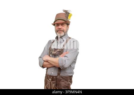 A bearded man wearing lederhosen, a feathered hat, and a gray shirt stands with crossed arms on a white background. Stock Photo