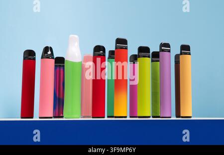 A range of colorful disposable vape pens lined up on a blue surface against a light blue background  Stock Photo