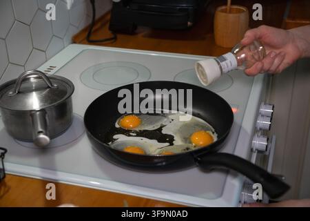 Hand seasons two fried eggs cooking in black pan on electric stovetop beside small saucepan. Home cooking, breakfast preparation, simple meal, kitchen Stock Photo