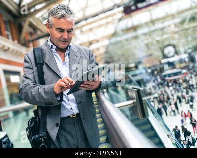 iPad Businessman using smart tablet travel busy commuter commuting rail Waterloo Station railway concourse with clock, business on move.  London UK Stock Photo