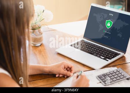 Woman writes notes at a home desk beside a laptop displaying a connected VPN and lock shield, illustrating secure remote work, privacy and data protec Stock Photo
