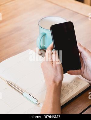 Close-up of hands holding a smartphone with a blank black screen above an open planner, pen and turquoise mug on a wooden table in a home workspace. Stock Photo