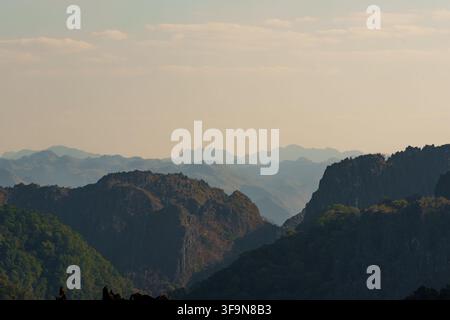 Forested karst ridges frame a deep mountain valley, with increasingly distant peaks fading into atmospheric haze beneath a broad pale sky. Stock Photo
