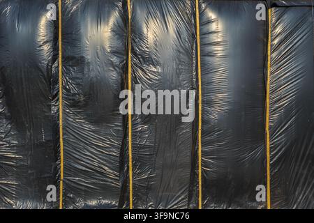 Full-frame abstract photograph features a textured black plastic film, secured by vertical yellow wooden planks. Stock Photo