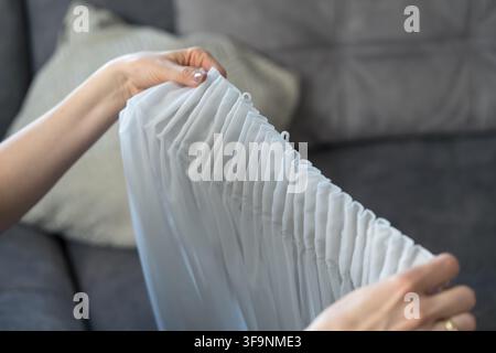 A woman's hands are inserting white plastic hooks into a white, semi-sheer curtain (sheer drape). Stock Photo