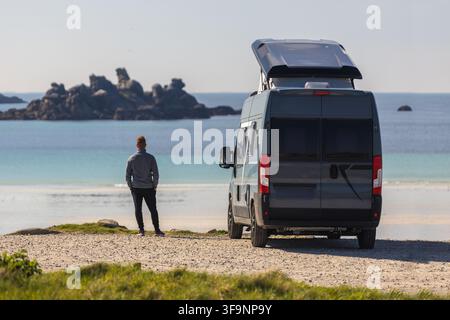 View of a man standing next to a grey camper van with a pop-up roof parked on a gravel path overlooking a sandy beach and rocky coastline in Normandy, Stock Photo