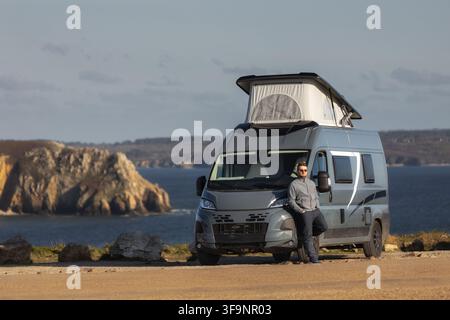 View of a man leaning against a grey camper van with a pop-up roof parked on the cliffside overlooking the sea and the rocky cliffs of Pen Hir in Cama Stock Photo