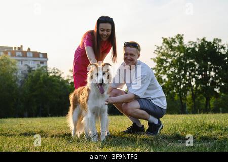 Happy couple posing with their dog on green grass in the park, enjoying leisure time on a sunny day. Stock Photo