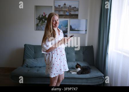 Smiling woman browsing smartphone while relaxing with her cat on couch in a cozy living room. Stock Photo