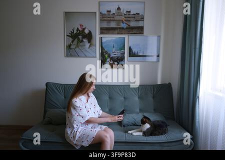 Young woman taking photos of her pet cat with smartphone while sitting together on sofa at home. Stock Photo