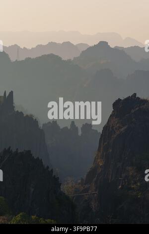 Jagged karst peaks and forested ridges recede through warm atmospheric haze, with a narrow suspension bridge crossing between rocky formations below. Stock Photo
