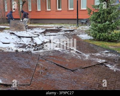 Two workers in hooded jackets are installing or removing large metal road plates on a muddy, snow-dusted ground surface. The heavy steel plates, Stock Photo