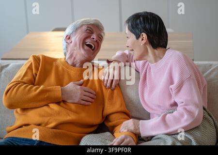 Funny cheerful senior husband and wife joking and sharing stories while sitting on couch together. Stock Photo