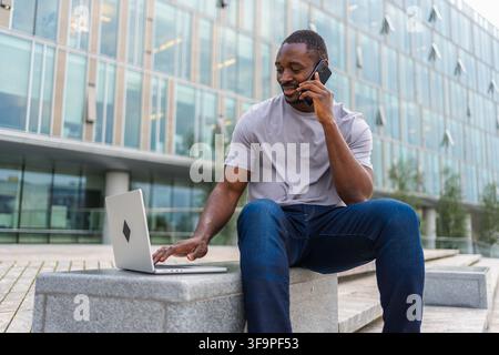 African American man freelancer using laptop talking on phone on urban street in city. Business man having virtual meeting. Professional programmer using phone laptop working outdoors. Remote work Stock Photo