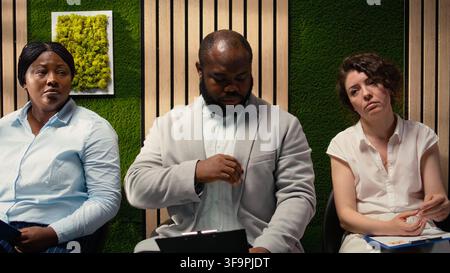 Diverse job seekers sitting in queue on the hallway with CV files, preparing for formal interview with the HR team. People reflecting impatience and nervousness during head hunting. Camera A. Stock Photo